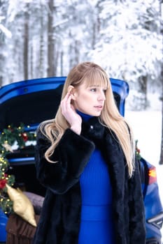 Woman in winter snowy forest in blue dress next to blue car decorated with Christmas decor. Christmas and winter holidays concept