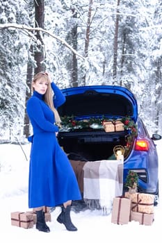 Woman in winter snowy forest in blue dress next to blue car decorated with Christmas decor. Christmas and winter holidays concept