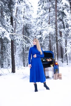 Woman in winter snowy forest in blue dress next to blue car decorated with Christmas decor. Christmas and winter holidays concept