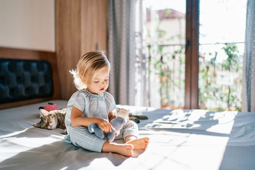 Little girl sits on a bed and combs a toy next to a lying cat. High quality photo