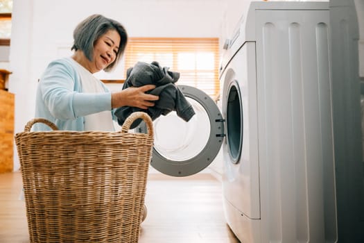 A happy senior woman, a smiling grandmother, loads dirty clothes into the washing machine, showcasing a solution for modern housework. Her routine reflects care and industry.