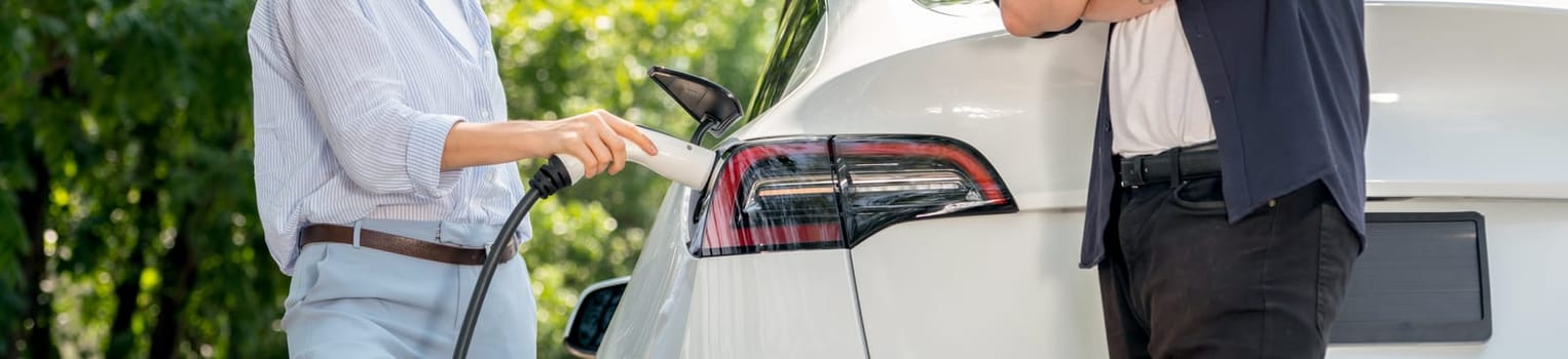 Focused EV car recharging electricity for battery on blurred background of lovey couple ruing their road trip travel by eco friendly electric car in national park and greenery forest on holiday. Exalt