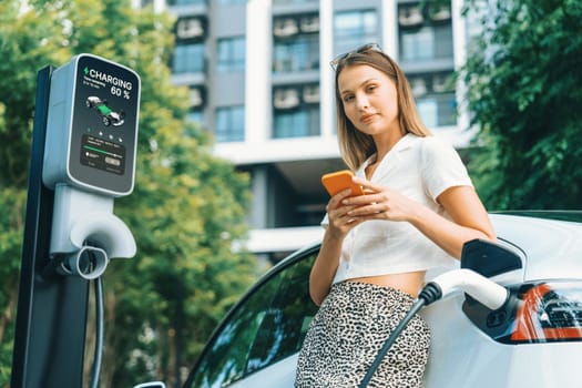 Young woman travel with EV electric car charging in green sustainable city outdoor garden in summer. Urban sustainability lifestyle by green clean rechargeable energy of electric BEV vehicle innards