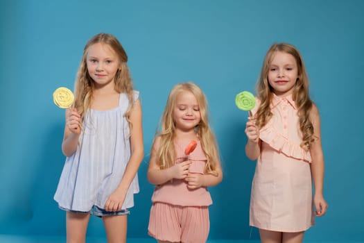 Three fashionable little girls in light dresses eat candy lollipop