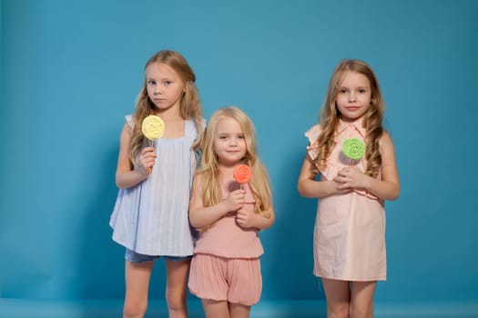 Three fashionable little girls in light dresses eat candy lollipop