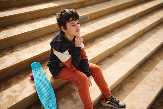 Thoughtful multi ethnic teenager, schoolboy in sportswear, sitting with his skateboard on the steps outdoors, dreamily looking into the distance. People. Active lifestyle. Sports Recreation.