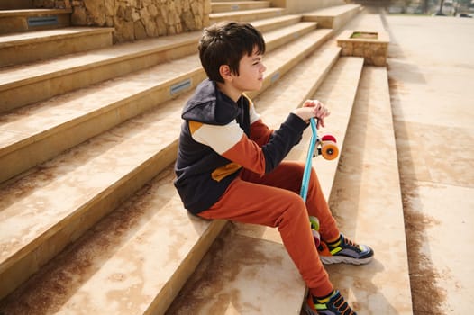 Portrait of a teenage student boy, schoolboy in sportswear sitting on steps in the urban skatepark, thoughtfully looking into the distance. Summer skateboarding, active lifestyle. Sports recreation