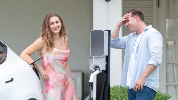 Happy and lovely couple with eco-friendly conscious recharging electric vehicle from EV home charging station. EV car technology utilized for residential home to future sustainability. Synchronos