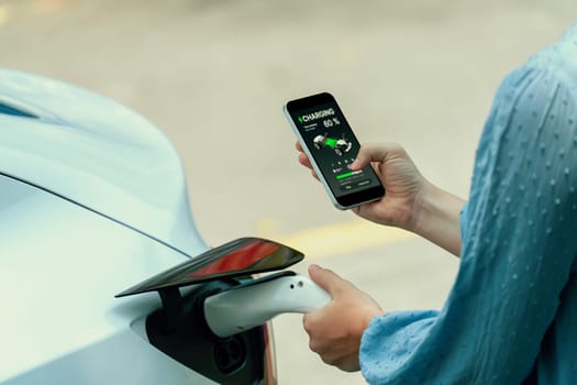 Hand insert EV charger plug into electric vehicle to recharge EV car, battery status display on smartphone EV application. Future alternative clean and sustainable energy for transportation. Perpetual