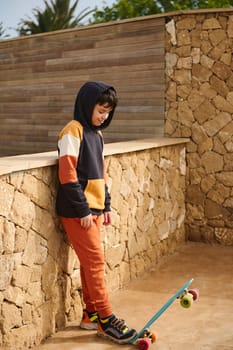 Full length portrait of happy child boy in stylish sports clothes, putting one leg on his penny board, smiling looking down, standing against a stone wall background outdoors. Leisure activity. People