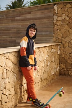 Full length portrait of handsome teenager boy in sportswear, playing with a skateboard, smiling at camera, standing outdoors against a stone wall background. People. Sport. Active healthy lifestyle