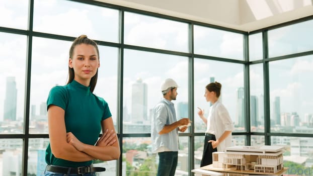 Young beautiful interior designer crossing arms while engineer team talking about house design. Skilled businesswoman smiling at camera with confident while sitting near house model. Tracery