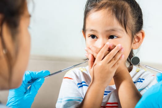 Dental kid examination. Doctor examines oral cavity of child uses mouth mirror to check teeth cavity but little girl cover mouth no need to checking she afraid, scared and closes his mouth with hands