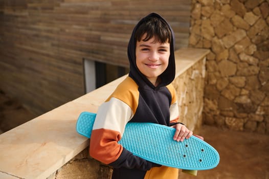 Lifestyle portrait of a teenager boy, hipster, skateboarder in trendy hoodie, holding his blue skateboard, smiling looking at camera, standing outdoors, against a stone wall background. Copy ad space
