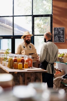 Caucasian man wearing a hat and holding boxes is interacting with male shopkeeper in grocery store filled with eco friendly and organic food products. Farmer carrying packages while talking to vendor.