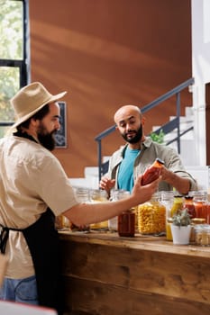 Salesman presenting organic items to vegan customer shopping for groceries, showing sauces and pantry goods in containers. Client thinking about buying fresh natural food produce.