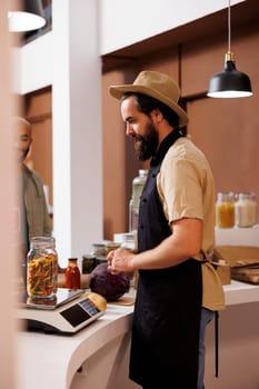 Male storekeeper, wearing a hat and apron, stands behind cashier desk while looking at jar filled with certain organic food item on measuring scale. Caucasian vendor weighing pasta for customer.