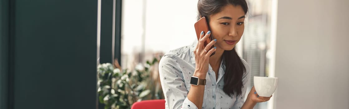 Busy female manager talking phone while working on laptop on modern office background