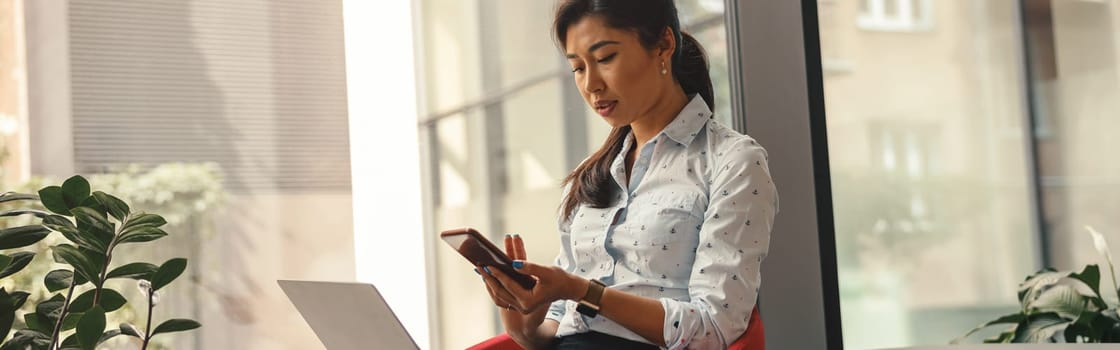 Woman entrepreneur looking on phone while working on laptop in office