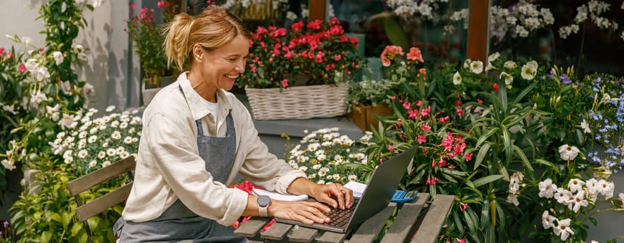 Small flower shop owner florist wearing apron working on laptop and taking online orders in store