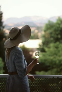 A woman, from behind, wearing a dress and a hat, stands with a wine glass in hand