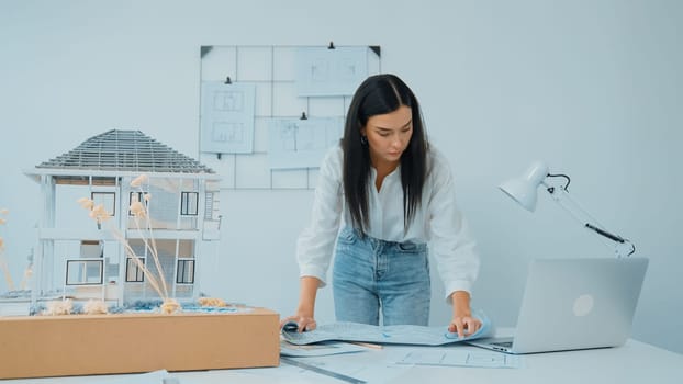 Young beautiful caucasian architect puts blueprint on the table and compare with house model while inspect hos model construction carefully at modern office with blueprint striped behind. Immaculate.