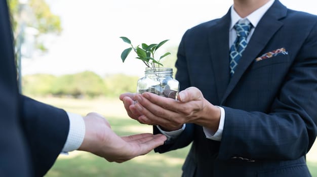 Concept of sustainable money growth investment with glass jar filled with money savings coins with businesspeople as eco-friendly financial investment nurtured with nature and healthy retirement. Gyre