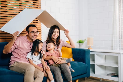 A joyful family during their home relocation sits on the sofa holding a cardboard roof mockup for protection. Safety happiness and planning for their new house is what makes them laugh.