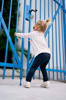 Little girl pulls the closed metal gates of the house towards herself. Side view. High quality photo