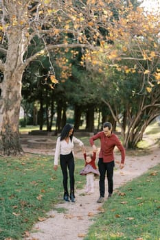 Mom and dad are walking along the path in the park, swinging their little daughter in their arms. High quality photo