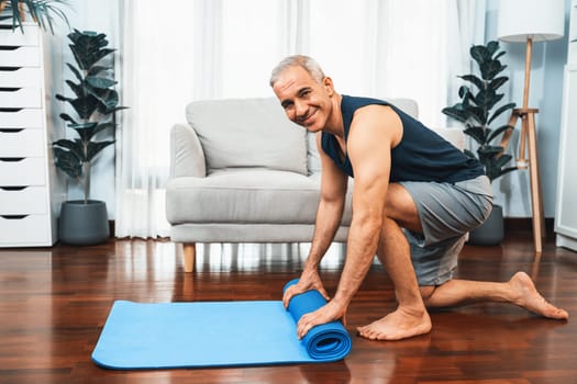 Active and sporty senior man preparing, rolling fitness exercising mat on living room floor at home. Home exercise as concept of healthy fit body lifestyle after retirement. Clout