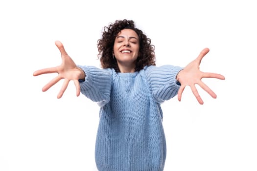 a young brunette woman with curls dressed in a blue knitted sweater stretches out her arms for a hug.