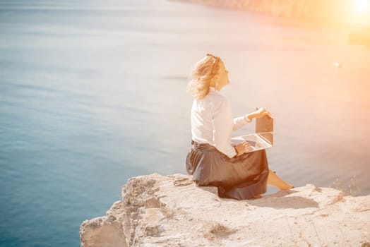 Business woman on nature in white shirt and black skirt. She works with an iPad in the open air with a beautiful view of the sea. The concept of remote work