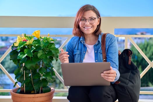 Positive smiling mature businesswoman with laptop outdoors. Middle-aged female in denim shirt headphones glasses with backpack looking at laptop screen. Work, business, technology, people 40s age