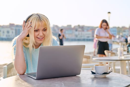 Smiling beautiful teenage girl in headphones looking into laptop. Blonde female in an outdoor cafe on waterfront using laptop. Youth, technology, lifestyle concept