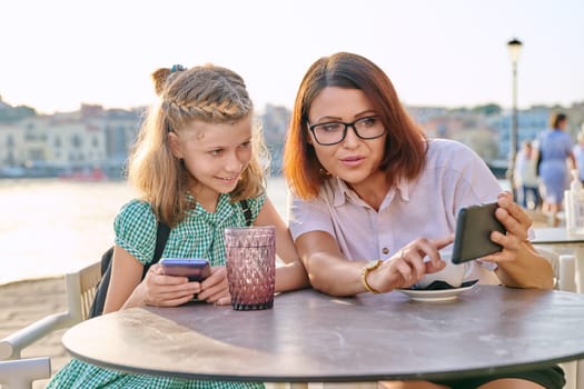 Mother and daughter child together in an outdoor cafe on the waterfront. Family, mom and preteen girl using smartphones, with coffee, drinks. Lifestyle, family, parent and child concept