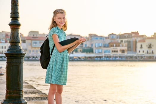 Girl child young student with a backpack reading a school notebook, outdoor, sunset sea embankment city background, copy space. Childhood, education, knowledge, back to school concept