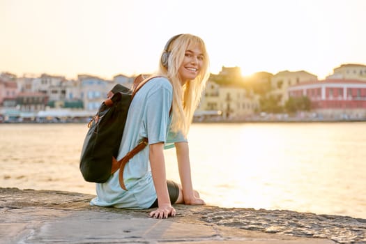 Happy having fun young teenage female in headphones with backpack enjoying old tourist European city, seascape, copy space. Tourism, Greece Crete Chania, summer, vacation, youth, rest, nature, sunset