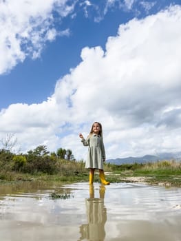 Little smiling girl in rubber boots stands in a puddle against a cloudy sky. High quality photo