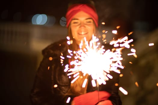 Woman holding sparkler night while celebrating Christmas outside. Dressed in a fur coat and a red headband. Blurred christmas decorations in the background. Selective focus.