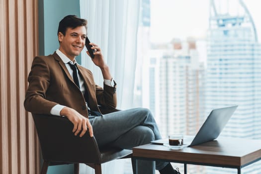 Closeup of handsome businessman making phone call with manager while sitting near window with skyscraper view. Executive manager talking working by using phone and laptop. Look aside. Ornamented.