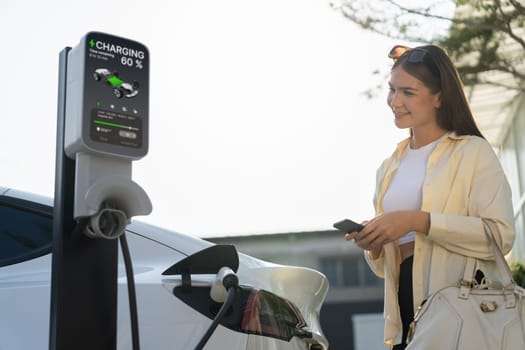 Young woman holding shopping bag and use smartphone to pay for electricity for recharging EV car battery from charging station at city mall parking lot. Modern woman go shopping by eco car. Expedient