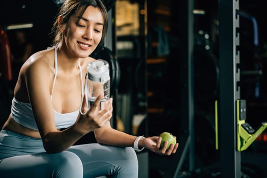 Smiling young woman holding a green apple and water bottle in fitness gym, emphasizing the importance of healthy food choices and hydration for achieving fitness goals. healthy lifestyle bodybuilding