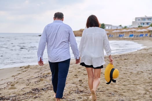 Happy middle aged couple walking together on beach, back view. Mature man and woman holding hands walking along seashore. Relationship, lifestyle, vacation, tourism, sea nature, people concept