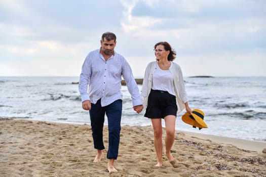 Happy middle-aged couple walking together on the beach. Mature man and woman holding hands walking along seashore. Relationship, lifestyle, vacation, tourism, sea nature, people concept