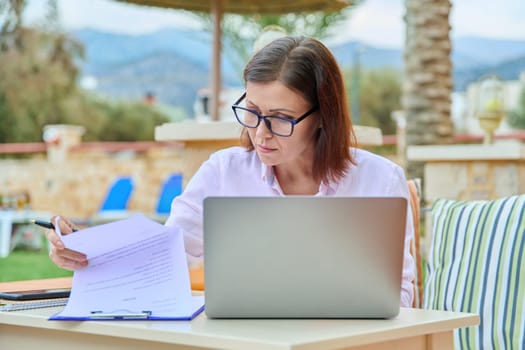 Middle aged woman working remotely using laptop. Female sitting at table writing, reading, outdoor hotel territory background. Summer season, freelance, distance learning and business