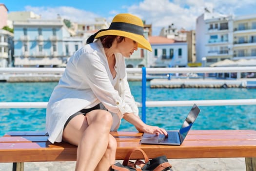 Mature woman in hat with laptop on promenade in city. Female freelancer tourist traveler typing text on laptop, sea harbor old city background. Tourism, summer, technology, business, travel, freelance