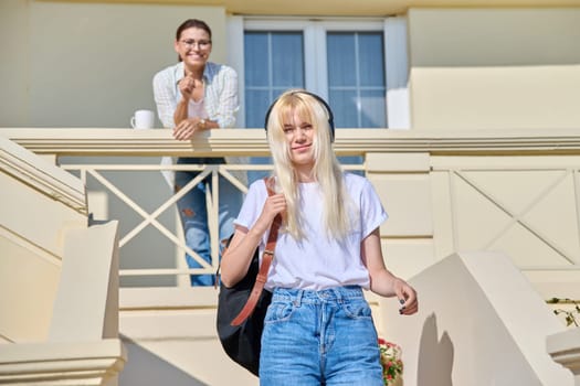 Middle-aged mother seeing off her teenage daughter on the porch of the house. Girl student in headphones with a backpack walking up the stairs. Family, lifestyle, parent and teenager concept