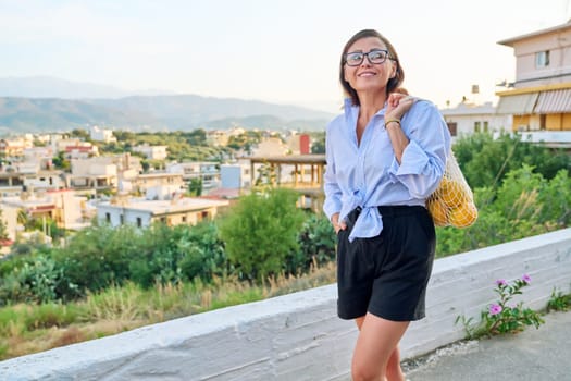 Eco tourism, woman walking relaxing in European mountain village, enjoying clean air, landscape, sunset, with a bag of fresh farm oranges. Nature, travel, ecology, lifestyle, people concept