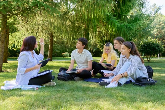 Outdoor, group of students with female teacher. Teenagers and mentor teacher talking sitting on grass in college park. Back to school, back to college, high school, education, teenagers concept